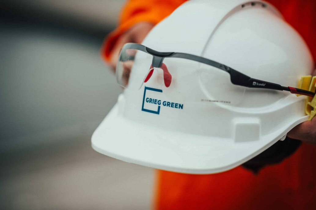 Grieg Green employee holds a white working helmet with the grieg green logo.
